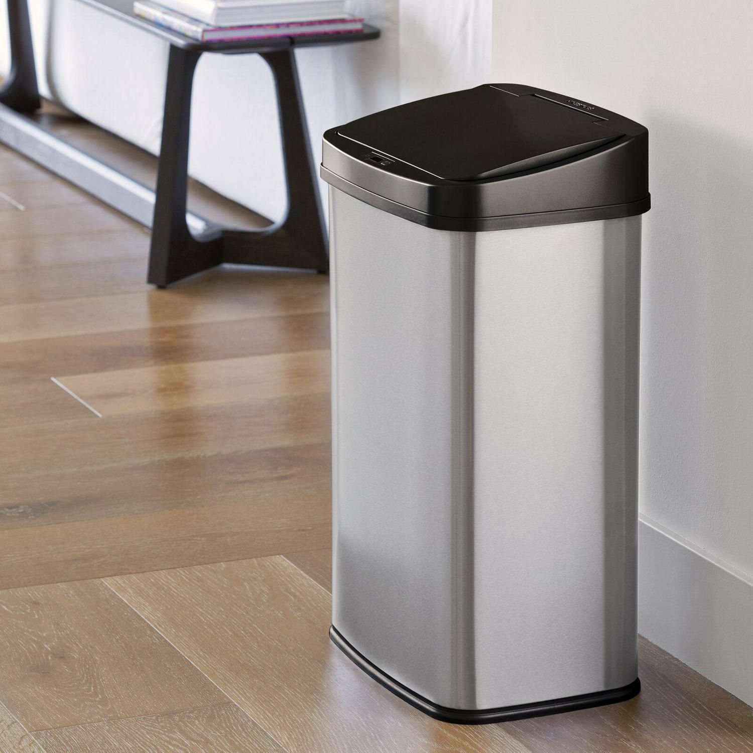 Stainless steel trash can with a black lid, standing on a wooden floor near a wall and a shelf.