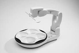 White robotic arm with a spoon dispensing food into a circular plate with four compartments.