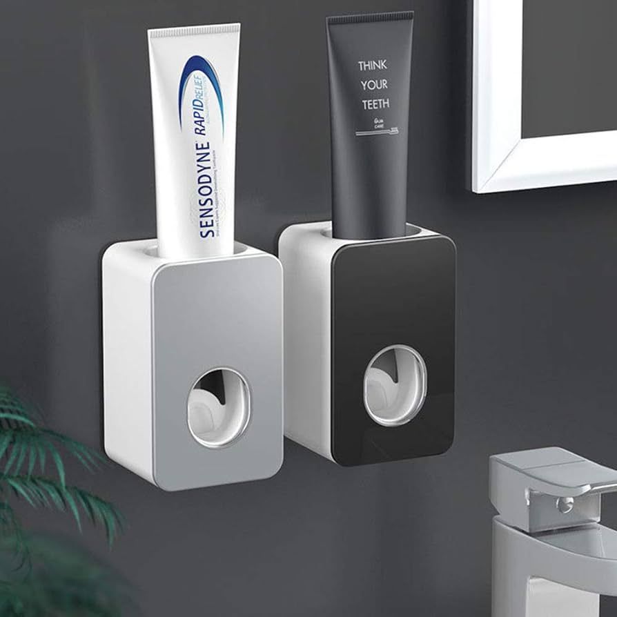 Two toothpaste dispensers, one silver and one black, on a grey wall, each with a tube of toothpaste.