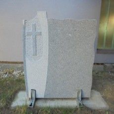 Ein Grabstein mit einem Kreuz darauf liegt im Gras vor einem Gebäude.