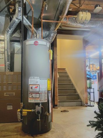 A water heater in a basement next to a furnace, stairs leading up, and exposed ductwork and pipes.