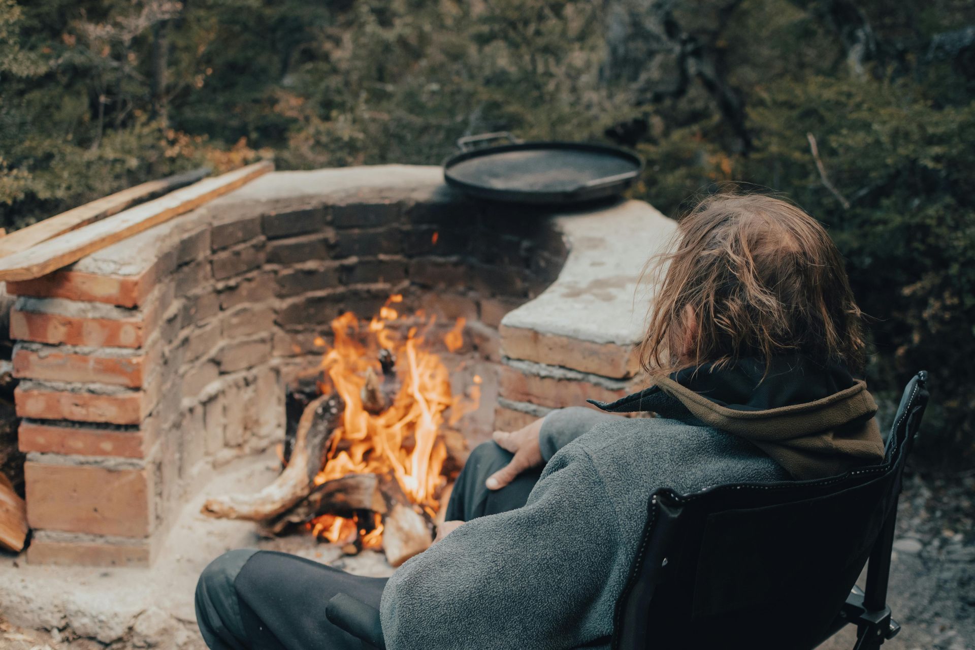Person sitting by a brick fire pit, with fire burning and pan above. Outdoors, in nature.