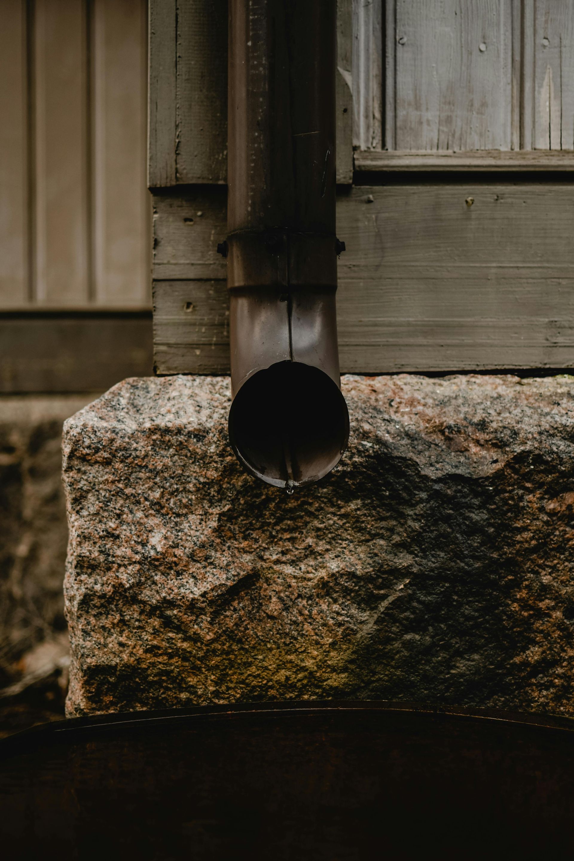 A dark brown gutter pipe draining onto a rough, textured stone foundation against a gray-green wall.