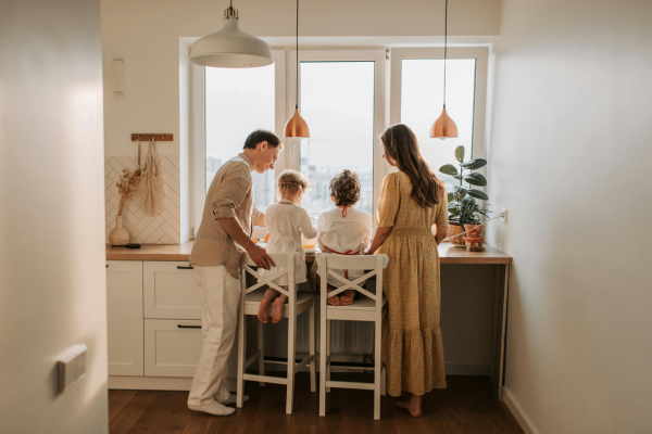 Family looking out a window in a kitchen. Two children sit on chairs, parents stand nearby. White cabinets, warm lighting.