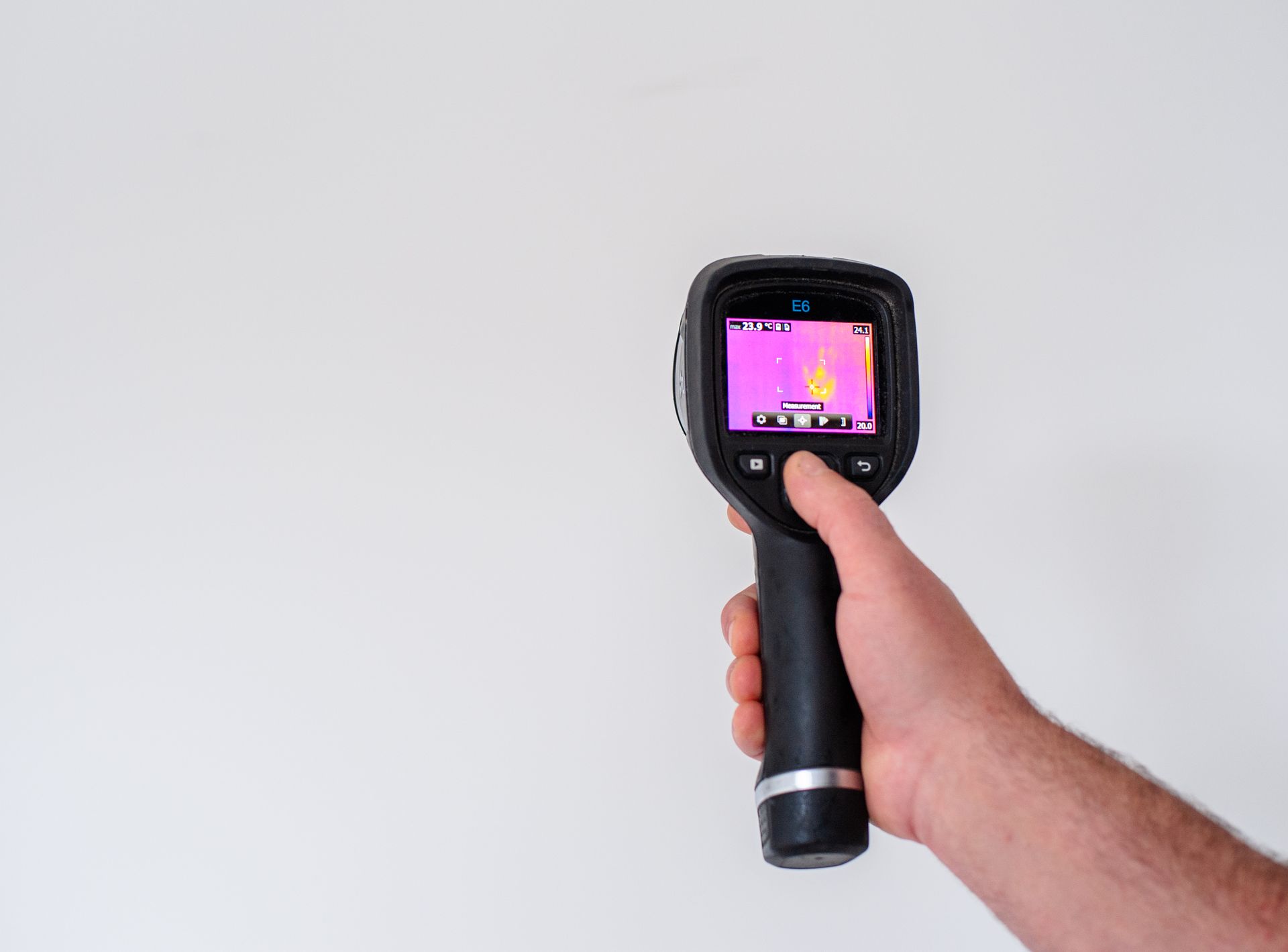 Hand holding a thermal imaging camera pointed at a white wall; display shows thermal variations in pink and yellow.