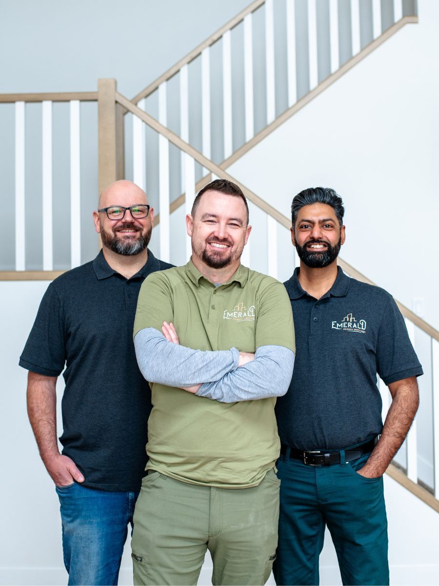 Three men smiling in front of a white staircase. Man in the center has arms crossed. All are wearing shirts with logos.