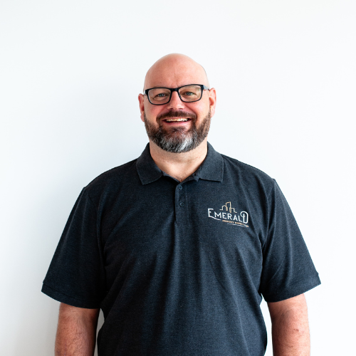 Ryan Watson smiles, wearing a dark gray polo shirt with “Emerald” logo. White background.