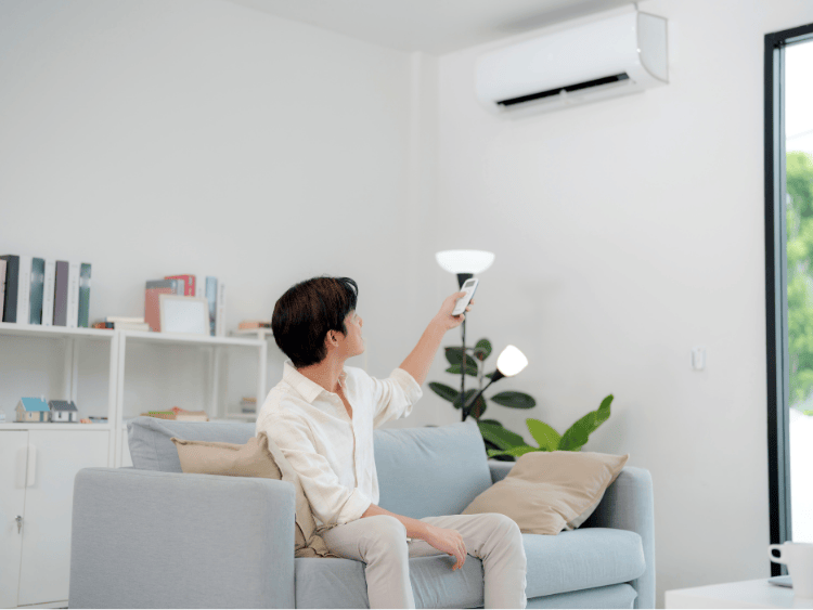 Person sitting on a couch points remote towards an air conditioner mounted on a white wall.