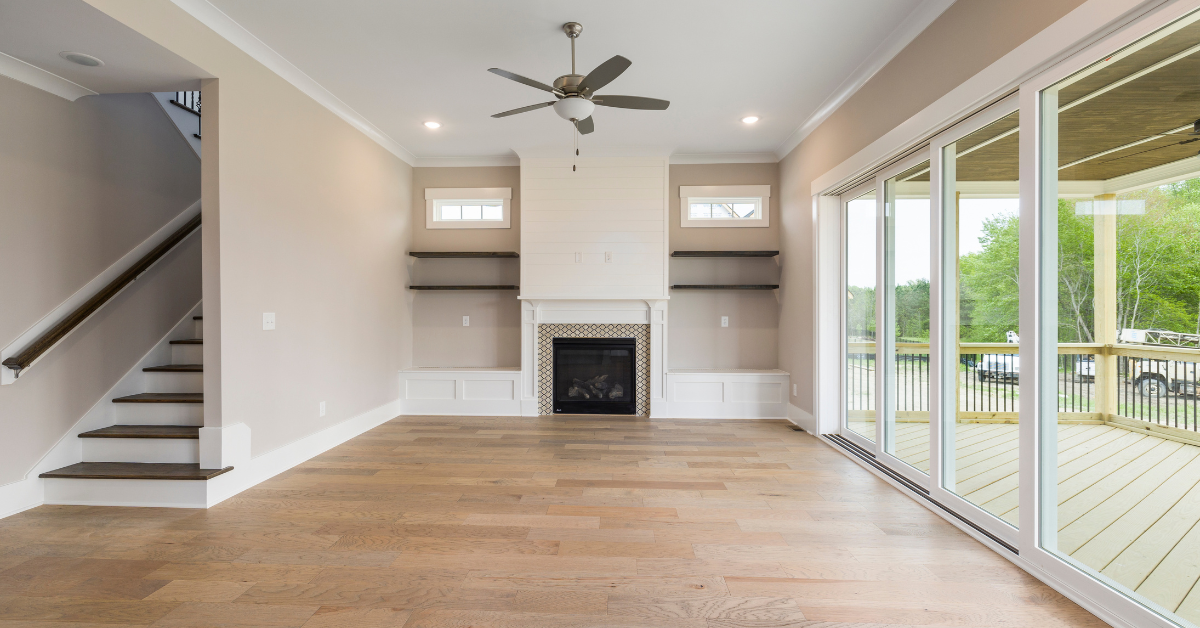 Empty living room with fireplace, stairs, and sliding glass doors to a deck; light wood floors.