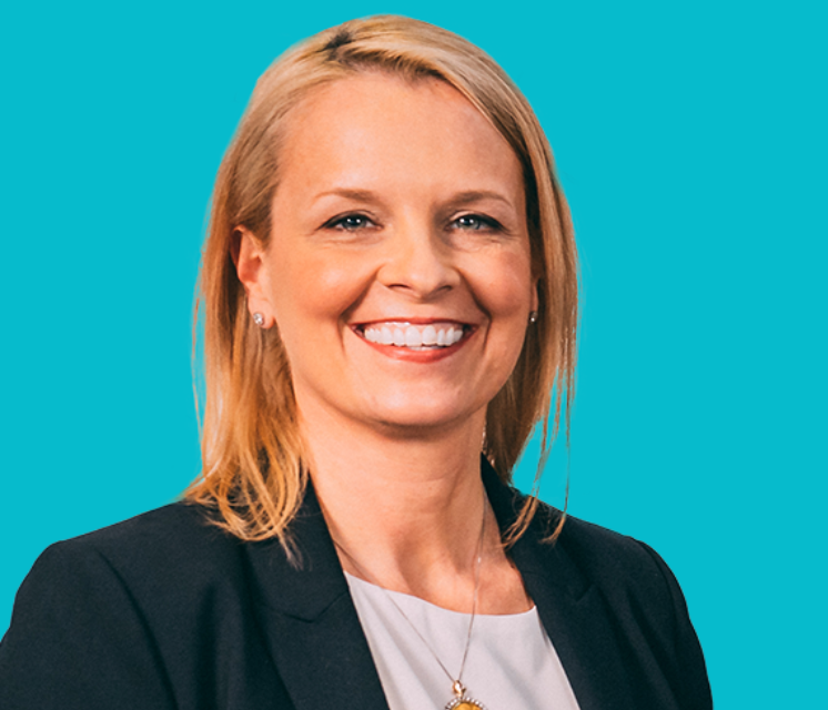 A woman in a suit and necklace is smiling in front of a blue background.