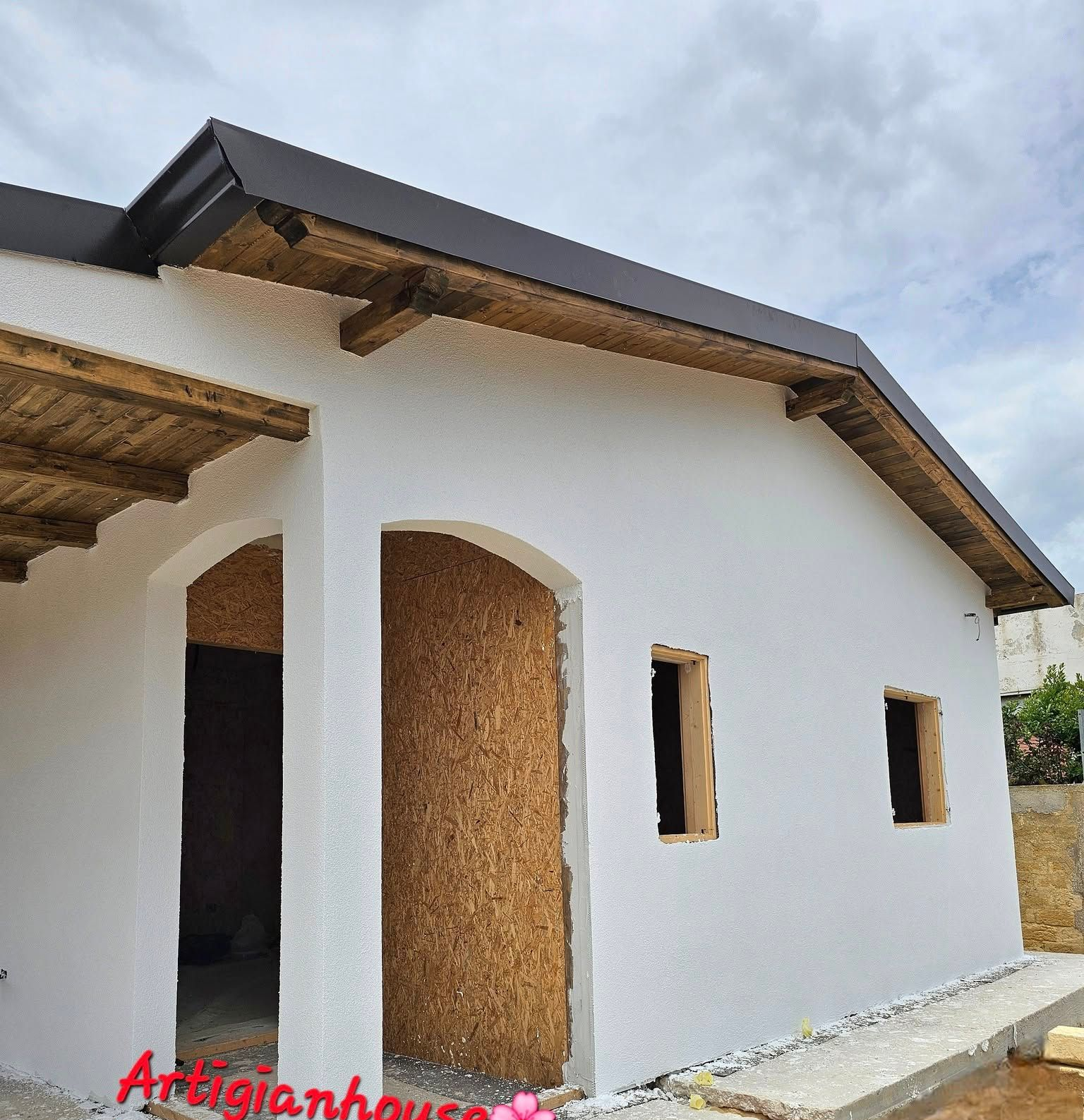 Facciata bianca di un edificio con finiture in legno, porta ad arco e piccole finestre sotto un cielo nuvoloso.