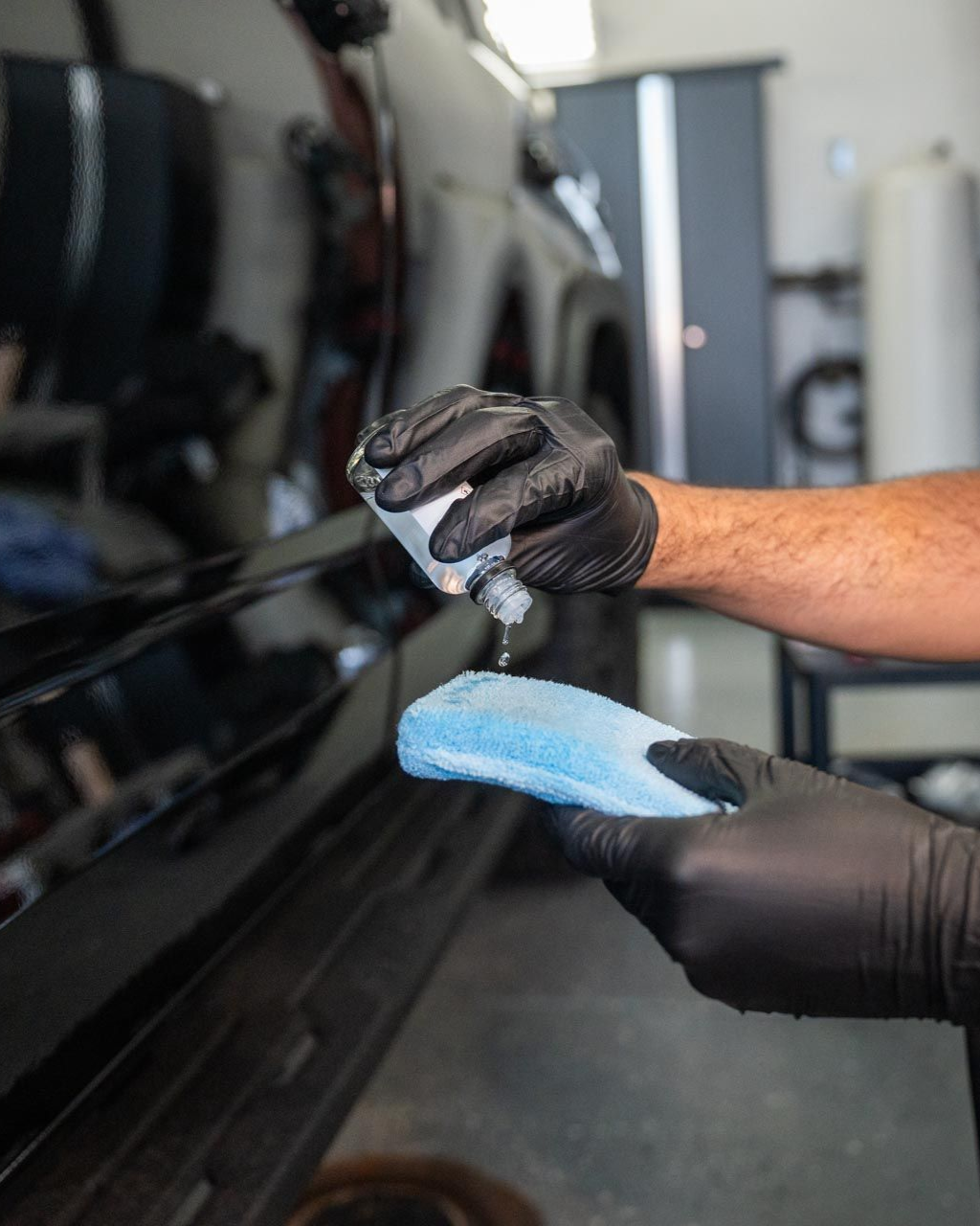 Person in black gloves applying liquid from a bottle onto a blue applicator pad and a black car.