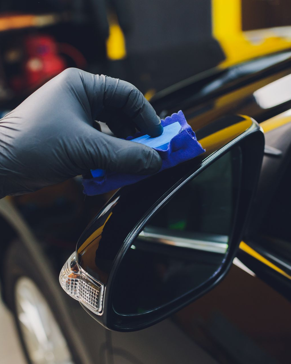 Gloved hand applying ceramic coating to a black car's side mirror with a blue applicator.