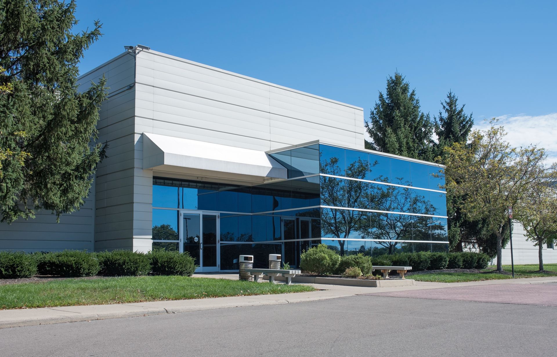 Modern building with blue glass entrance, awning, and trees on a sunny day.