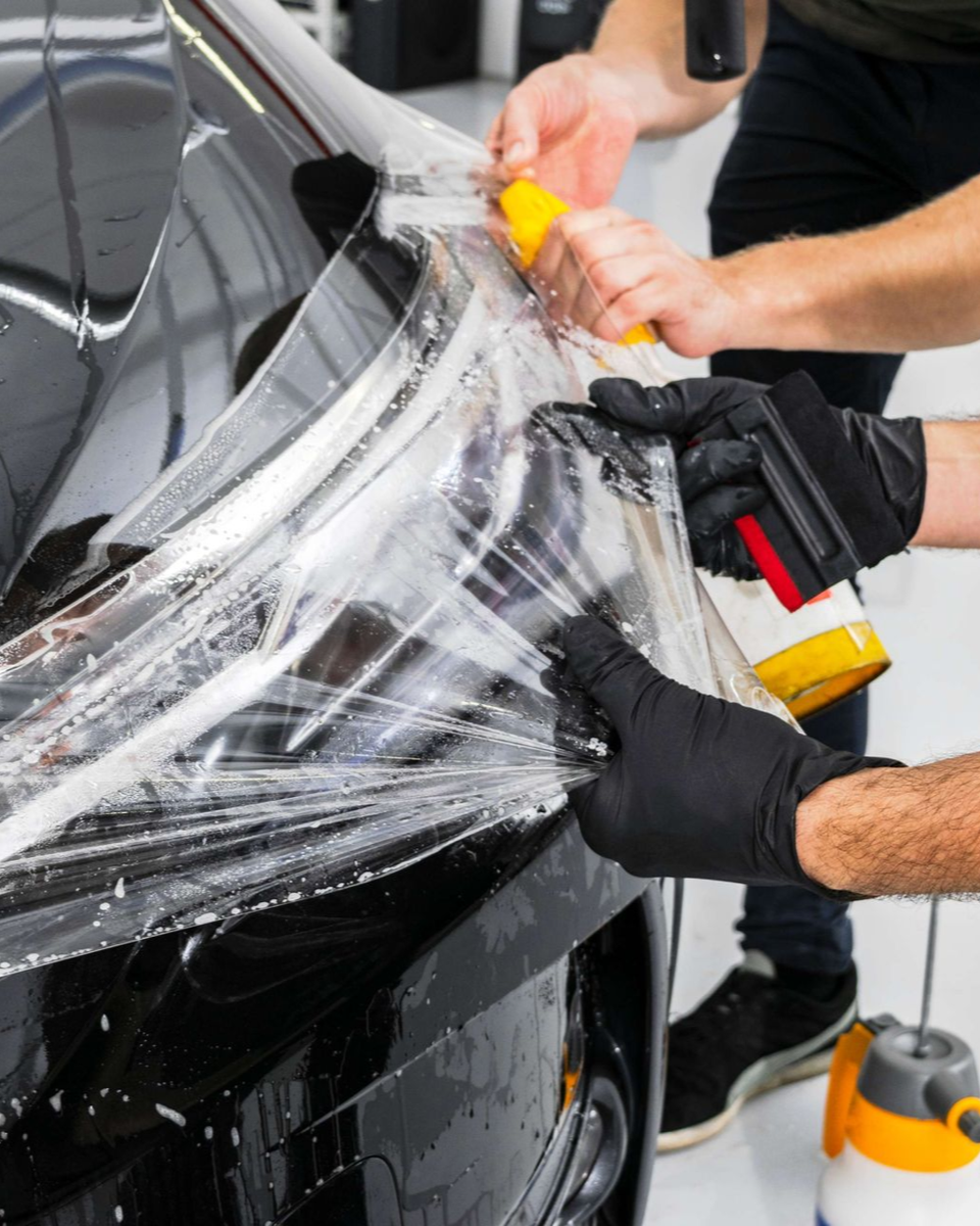 Two people installing clear protective film on a black car's bumper, using tools and spraying liquid.