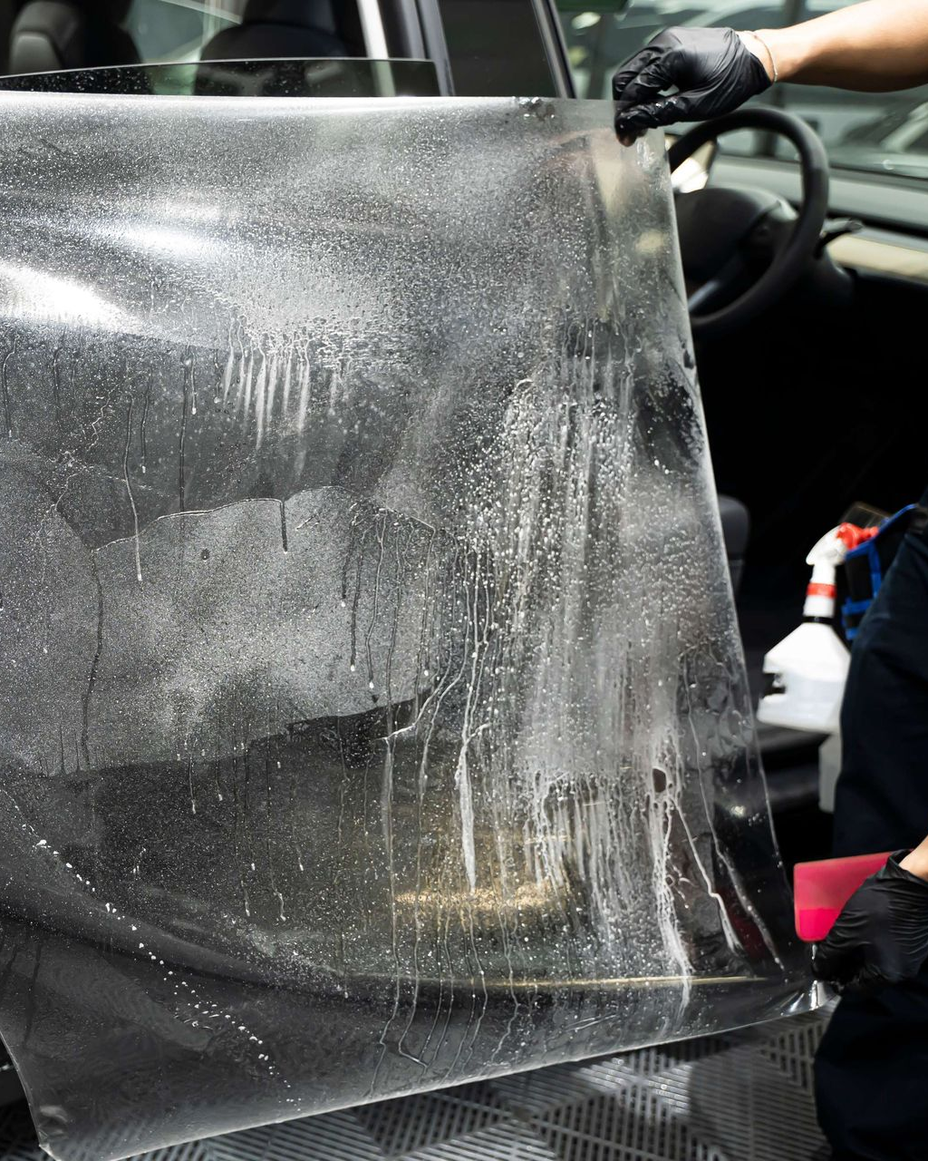 Person applying wet, clear film to a car window in a vehicle interior, wearing black gloves.