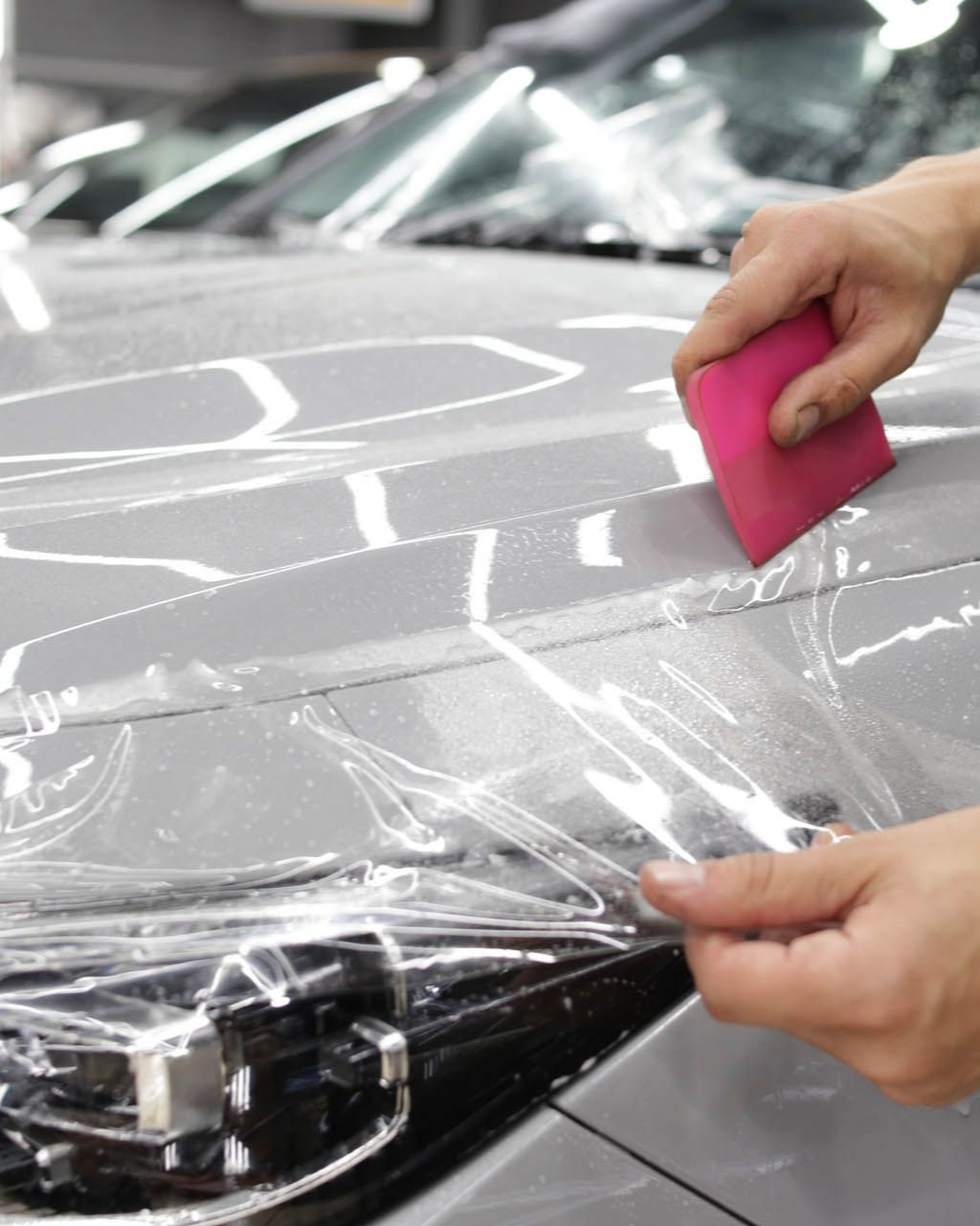 Hands applying clear protective film to a gray car hood with a pink smoothing tool.