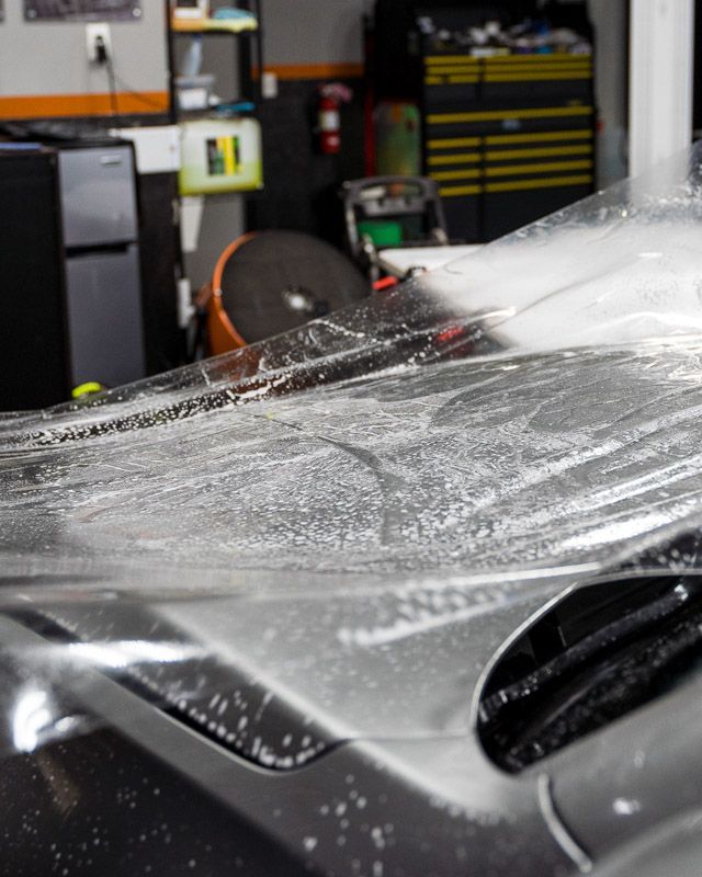 Clear film being applied to a car hood, showing bubbles and reflections in a garage.