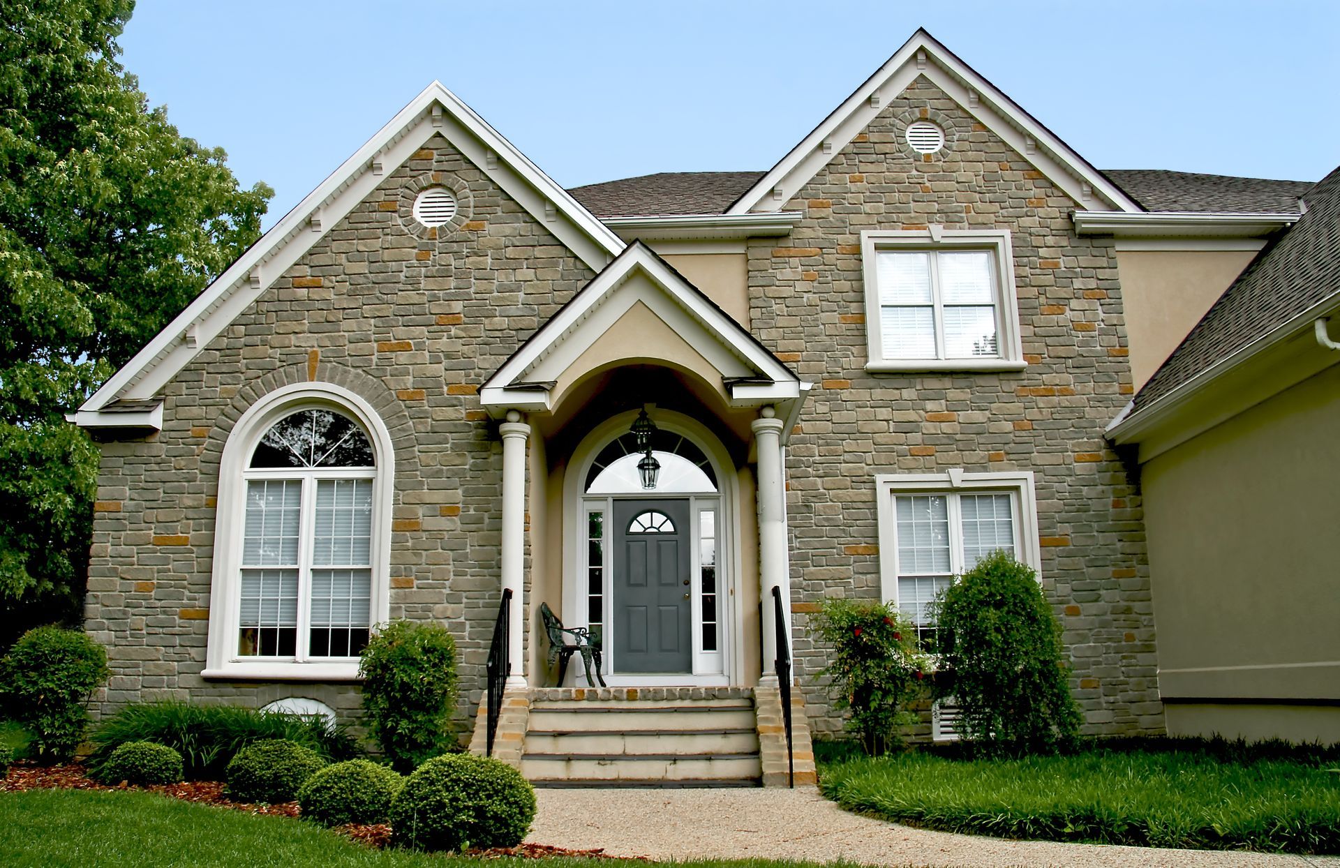 Stone-faced house with arched doorway, windows, and landscaping; gray door, light trim, green lawn, blue sky.