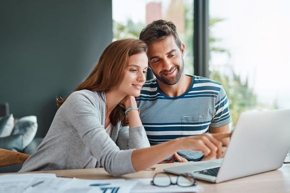 Two people smiling at a laptop on a table, reviewing something together in a bright room.