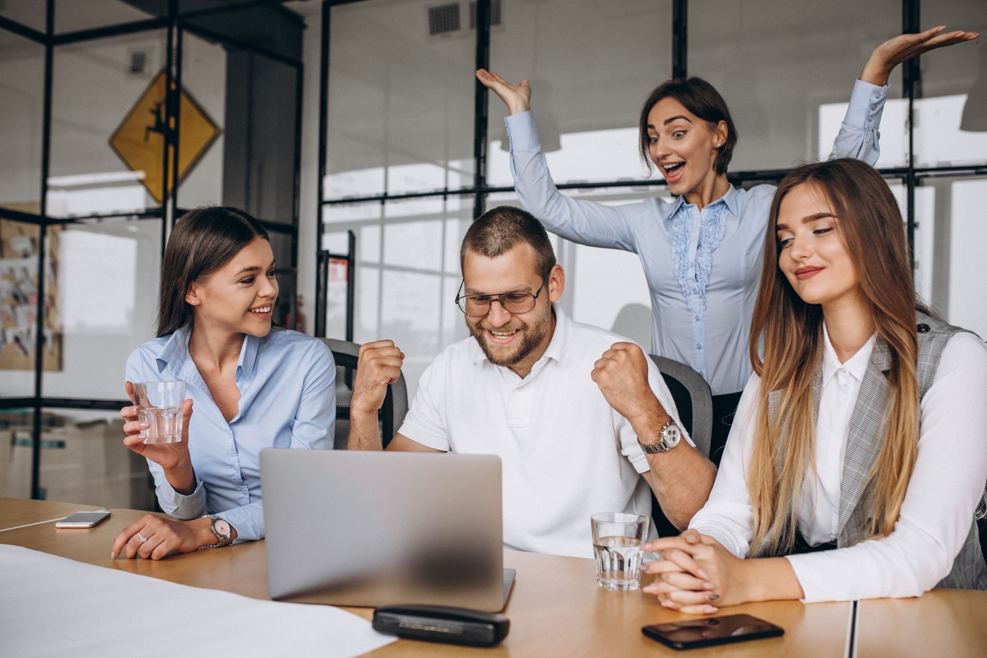 Coworkers celebrating around a laptop in a bright office, smiling and raising their arms