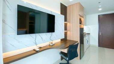 A modern studio apartment interior featuring a wall-mounted television, a wood desk, an office chair, and a kitchenette.