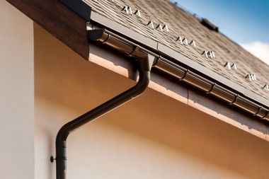 Brown downspout and gutter system attached to the side of a beige house with a shingled roof.
