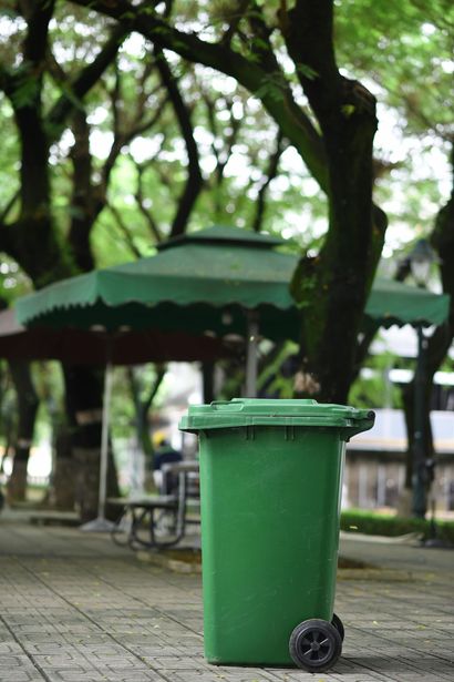 A green wheeled trash bin stands on a paved park path in front of a shaded seating area under large trees.