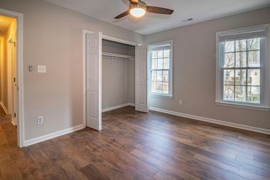 An empty bedroom with light walls, wood-look flooring, a white closet, and two windows with view of trees outside.