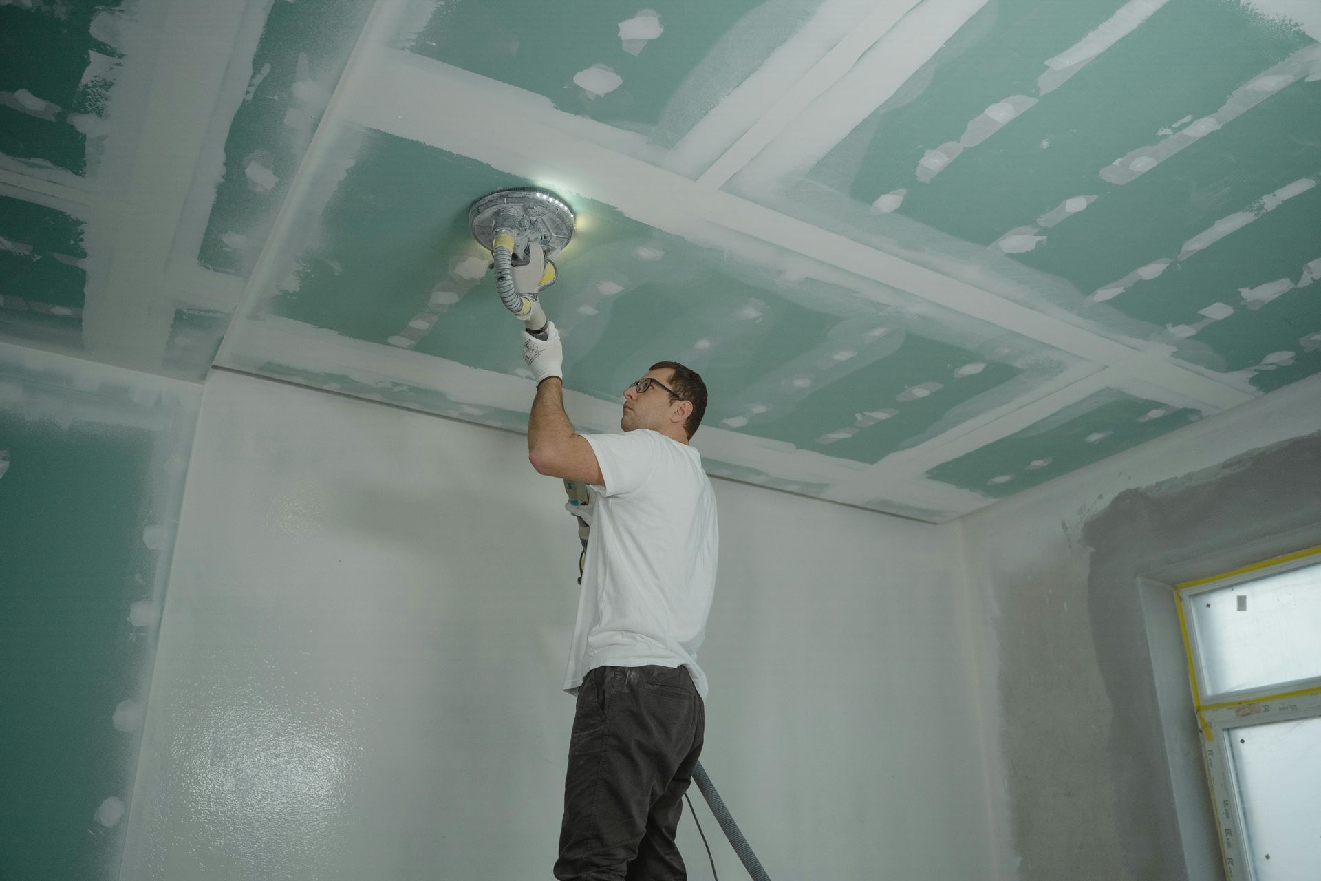 A person stands on a ladder, using a power sander to smooth the seams on a green drywall ceiling.