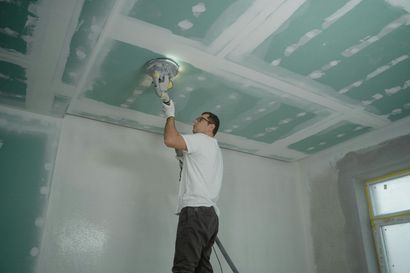 A person stands on a ladder, using a power sander to smooth the seams on a green drywall ceiling.