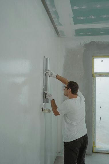 A person wearing gloves and safety glasses uses a metal trowel to smooth white plaster onto an interior wall.