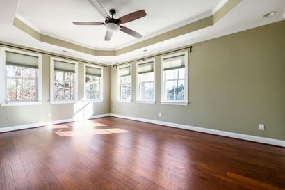 Empty living room with olive walls, dark wood floors, a tray ceiling with a fan, and several windows.