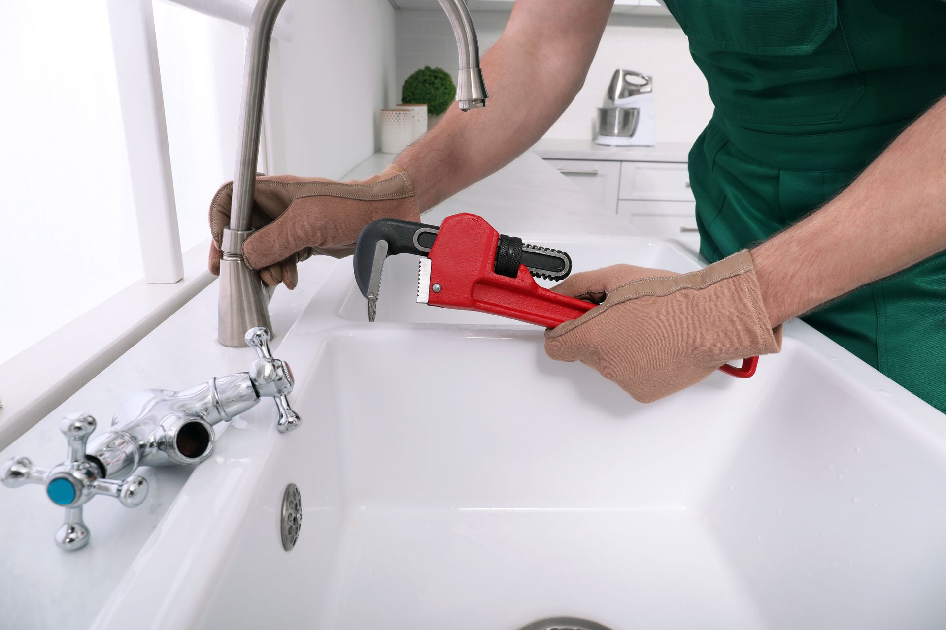 A person wearing work gloves and a green uniform uses a red pipe wrench to work on a bathroom sink faucet.