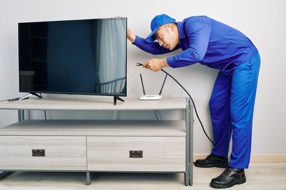 A technician in a blue uniform plugs a black cable into a television sitting on a wooden media console.