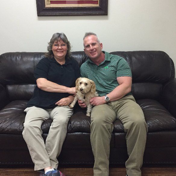 A man and a woman sit on a couch holding a small dog