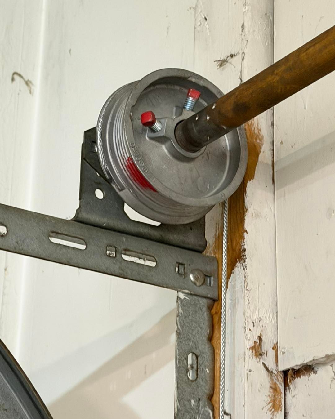 Garage door spring assembly mounted on a white wall, with a cable and wooden bar.