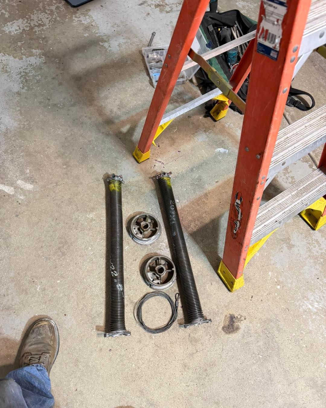 Two garage door springs, pulleys, and a cable lying on a concrete floor near a ladder.