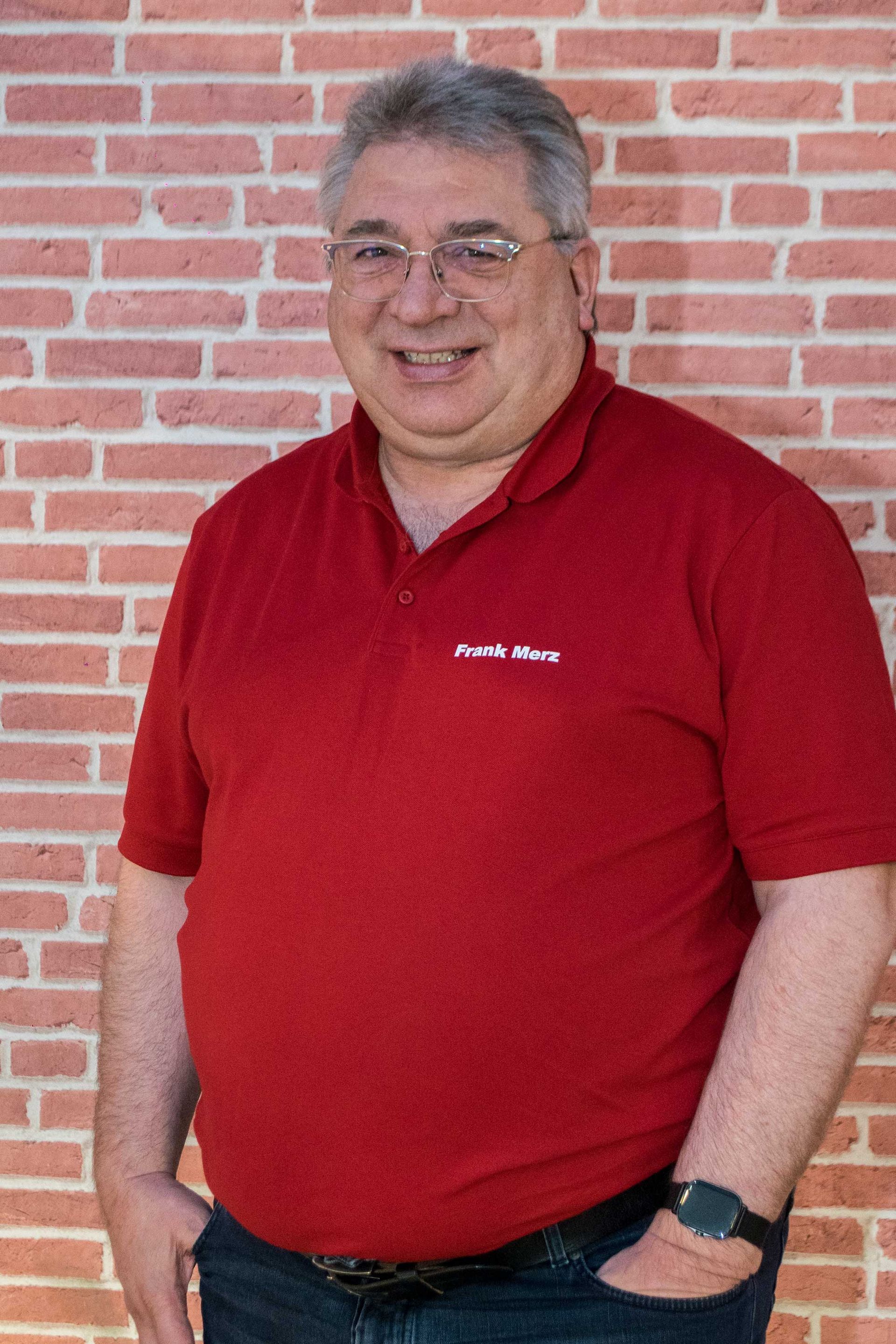 Frank Merz mit Brille und rotem Hemd steht vor einer Backsteinmauer.