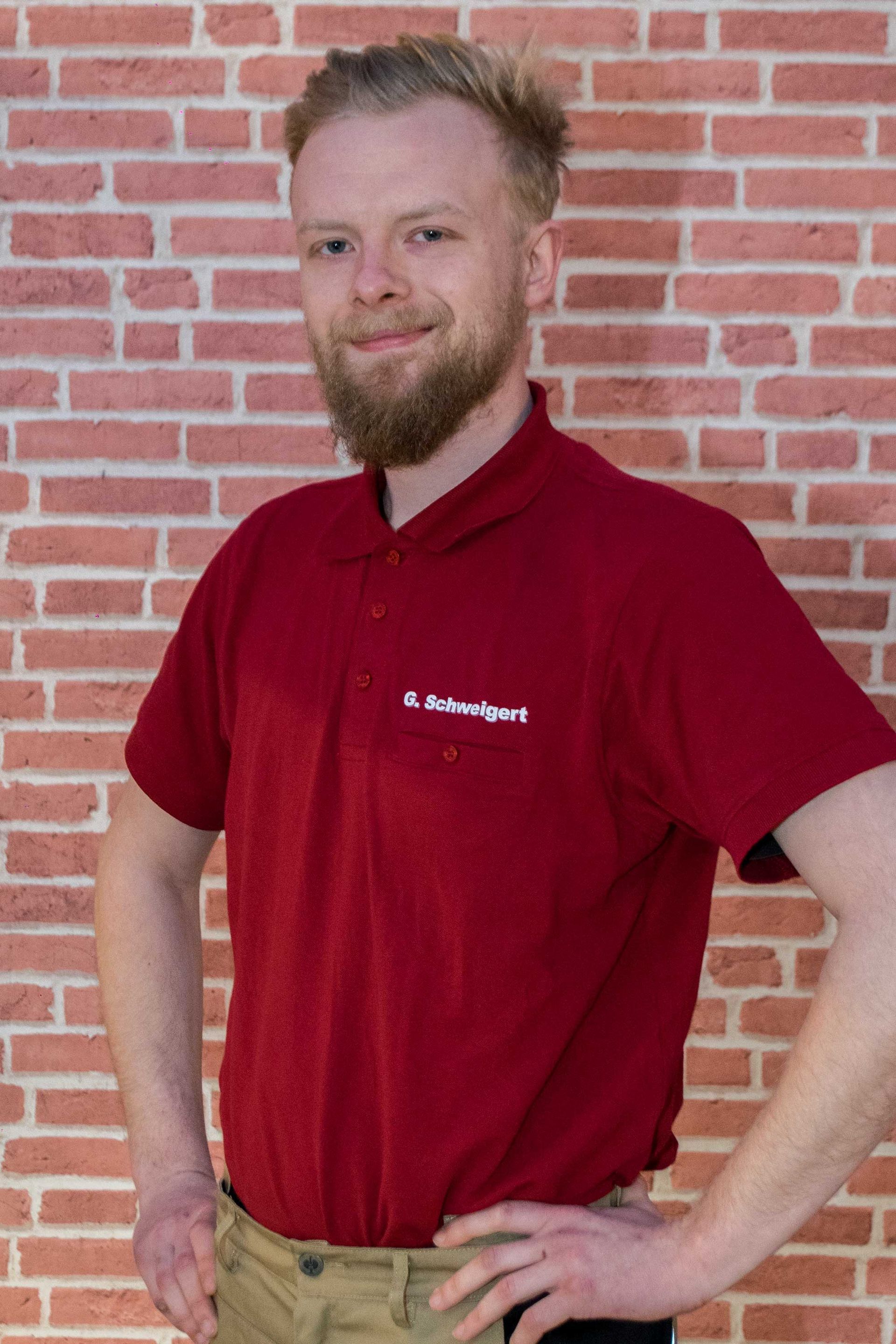 Gordian Schweigert mit Bart und einem roten Poloshirt steht vor einer Backsteinmauer.