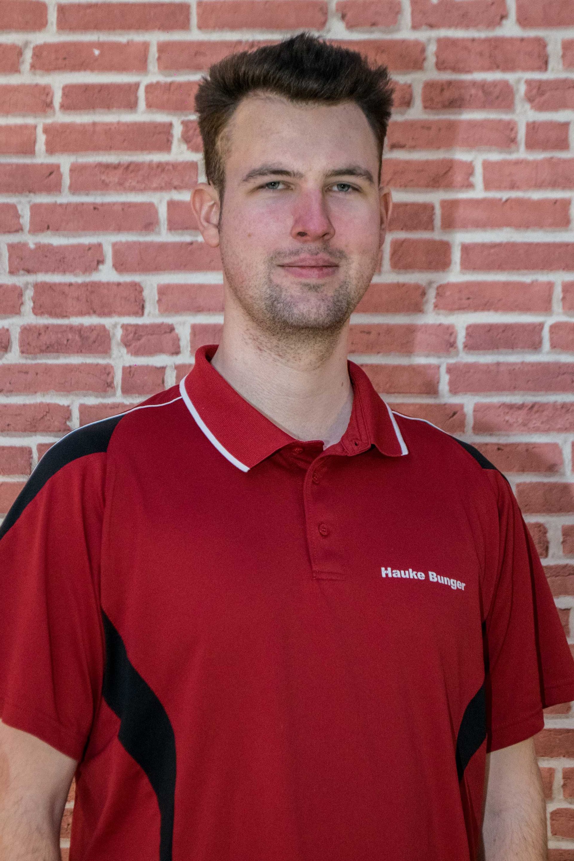 Hauke Bunger in einem roten Hemd steht vor einer Backsteinmauer.