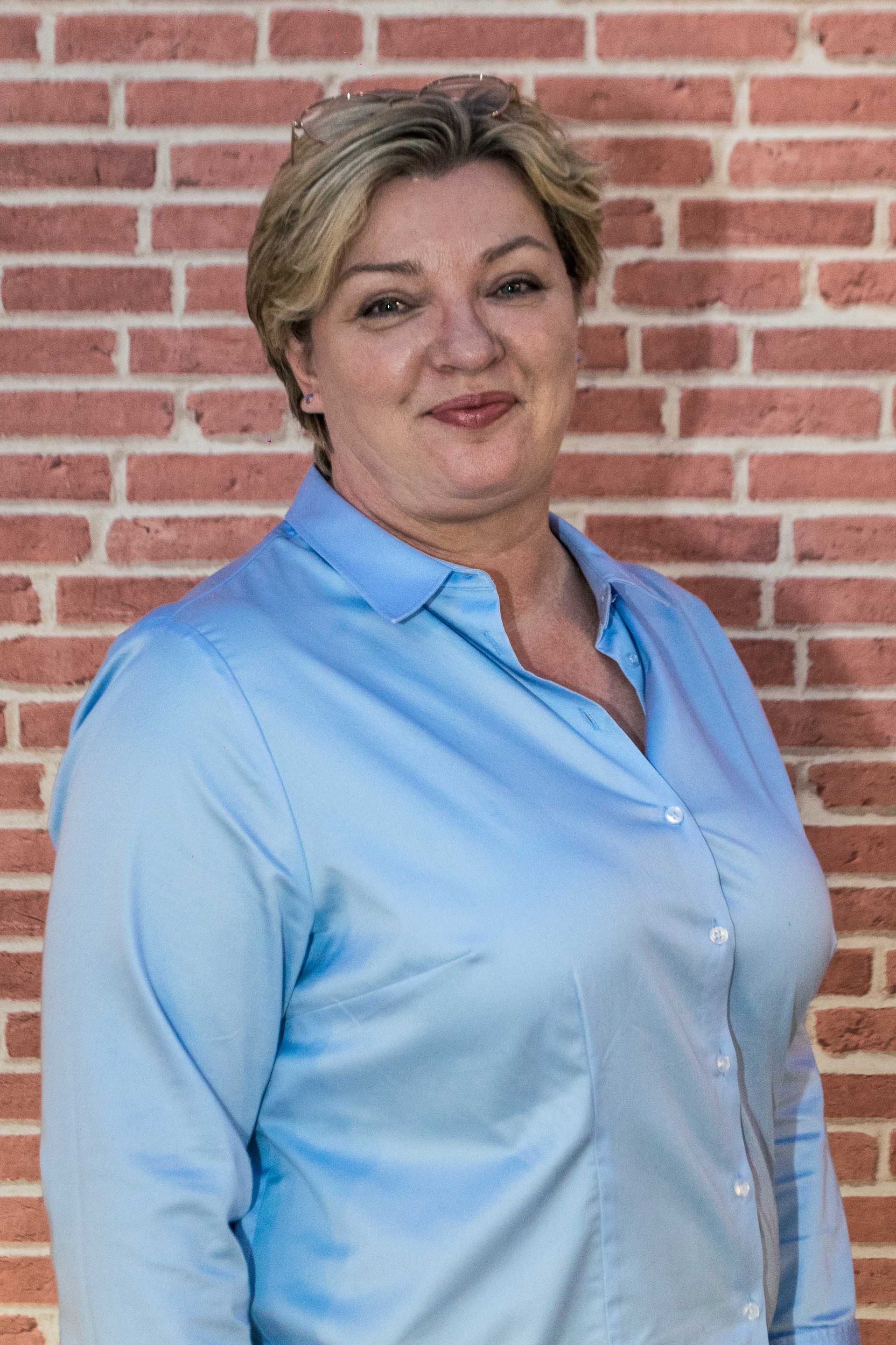 Heike Merz in einem blauen Hemd steht vor einer Backsteinmauer.