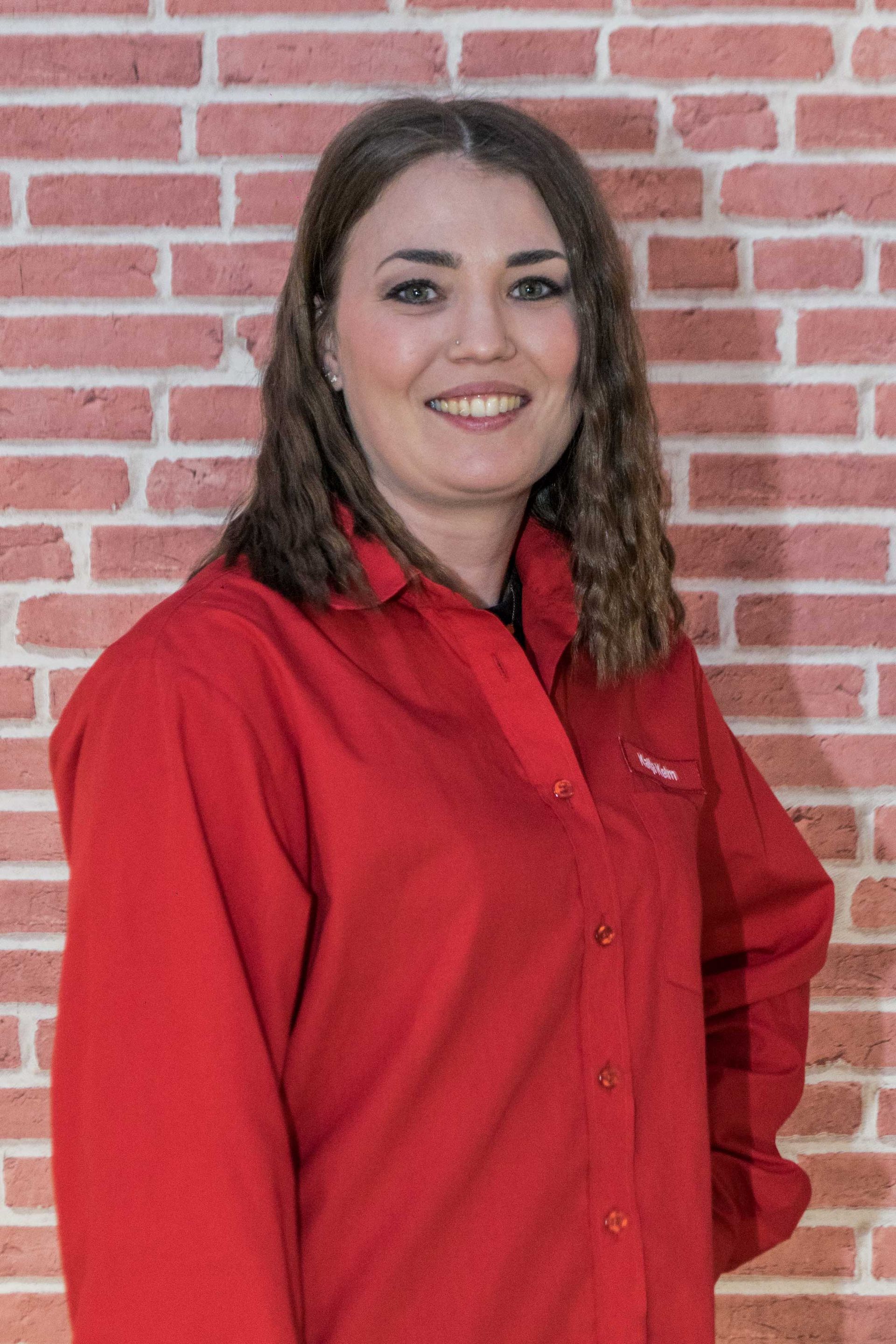 Katja Kelm in einem roten Hemd steht vor einer Backsteinmauer.