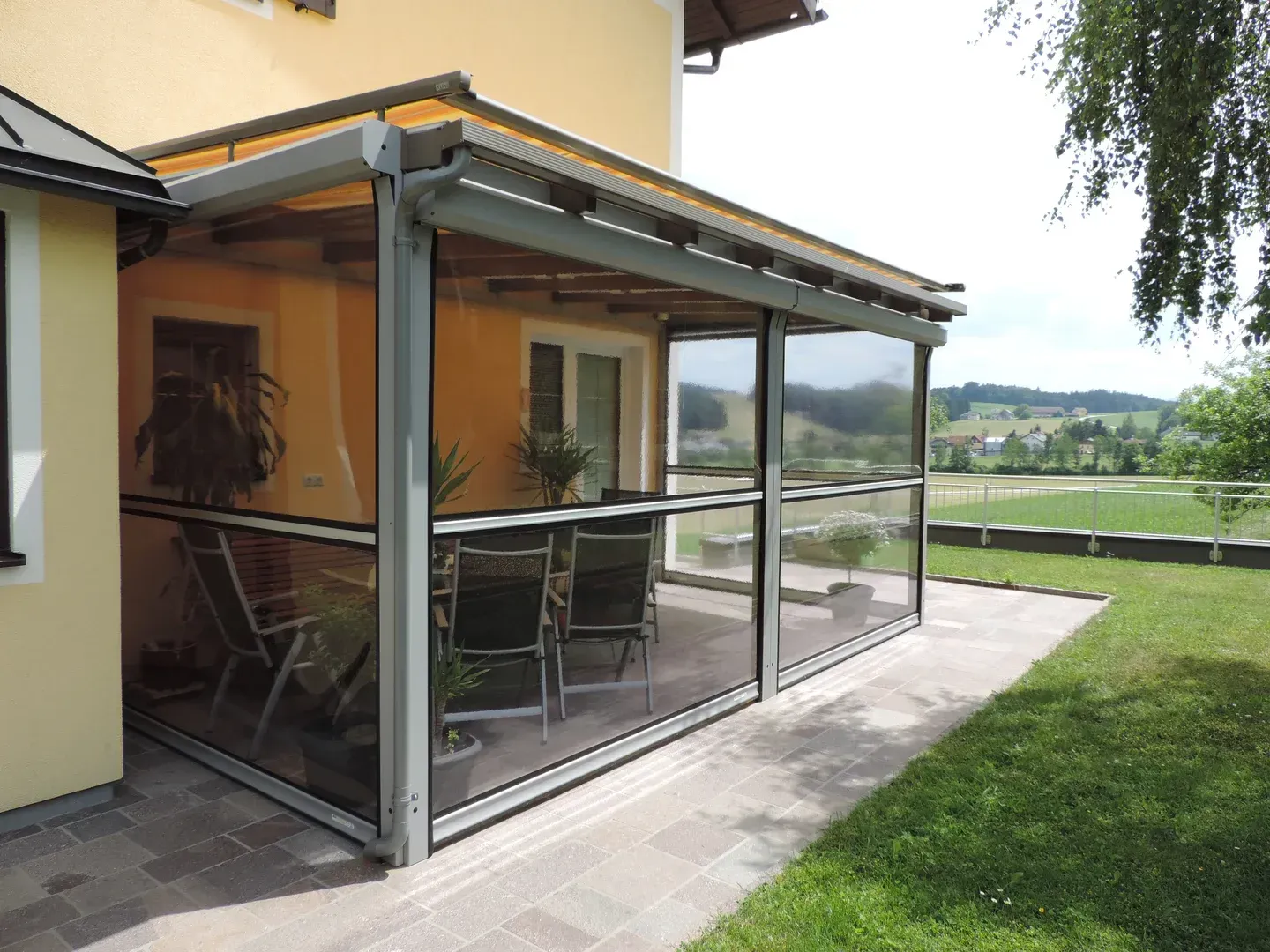 Rollfenster als transparenter Wind- und Wetterschutz an einer Terrassenüberdachung, seitlich geführt und teilweise geschlossen, mit freiem Blick in den Garten.