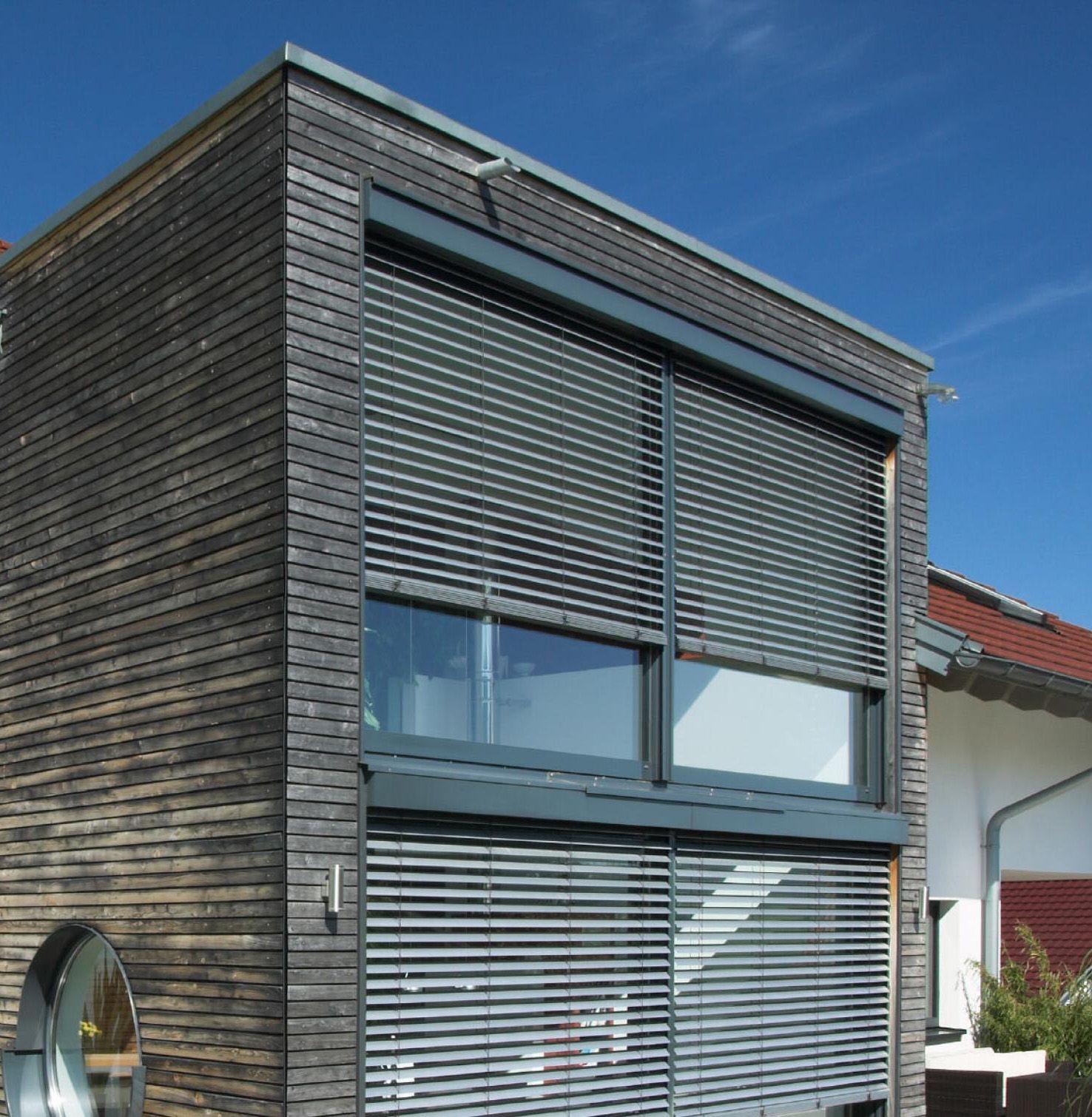 Außenansicht eines modernen Wohnhauses mit Holzfassade und großflächigen Fenstern, ausgestattet mit außenliegenden Raffstoren aus Aluminium zur flexiblen Lichtlenkung und wirksamem Sonnenschutz.