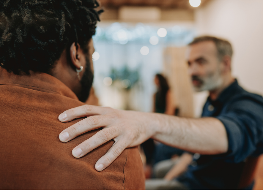 A person places a supportive hand on the shoulder of another person in a softly lit, group meeting setting. A person places a supportive hand on the shoulder of another person in a softly lit, group meeting setting.