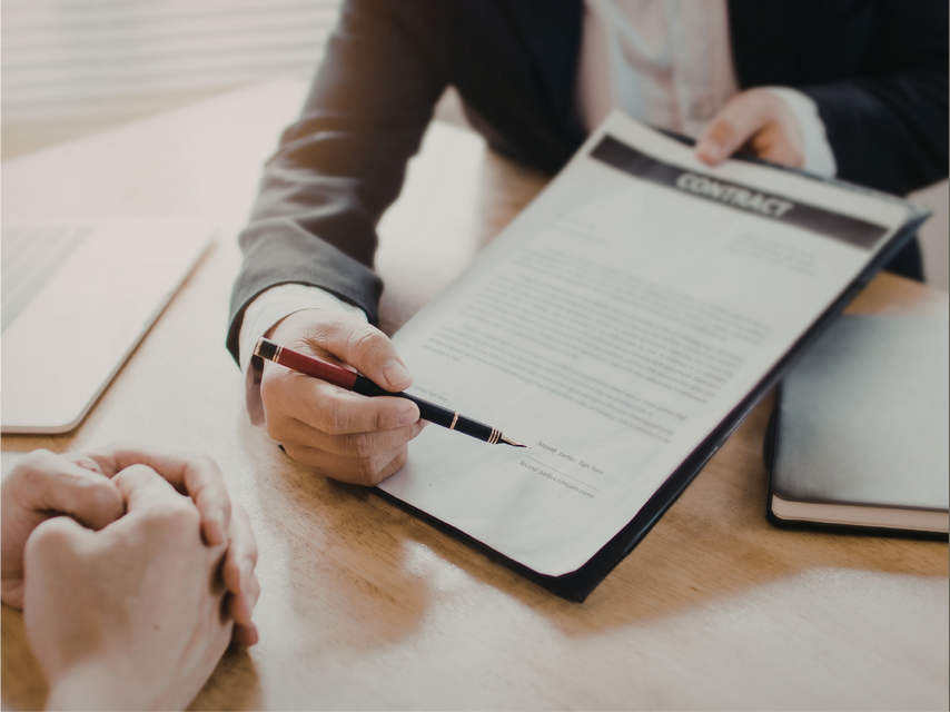 A person in a business suit points to a line on a contract, requesting a signature from someone sitting across the desk.