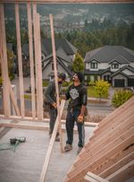 Two construction workers building a wooden frame on a rooftop overlooking houses and trees.