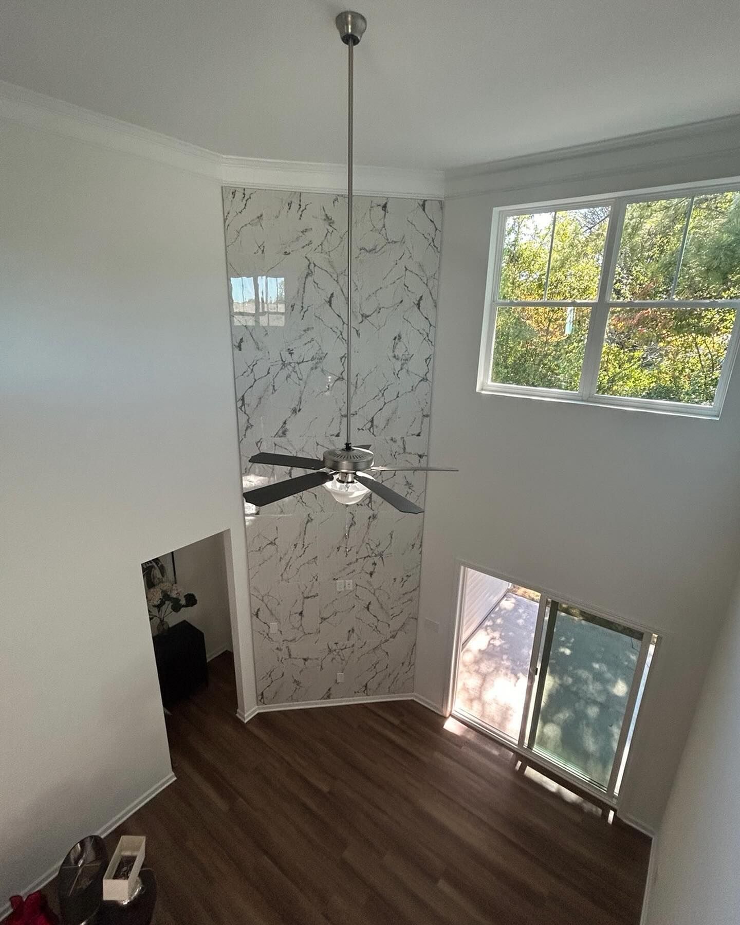 Two-story room with a ceiling fan. Marble-look wall with a window. Large window looking out to trees. Wooden floor.