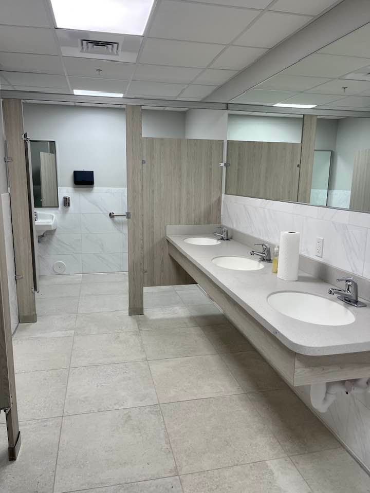 Public restroom with three sinks, mirrors, and stalls. Light-colored tile floor and wood-look stall doors.